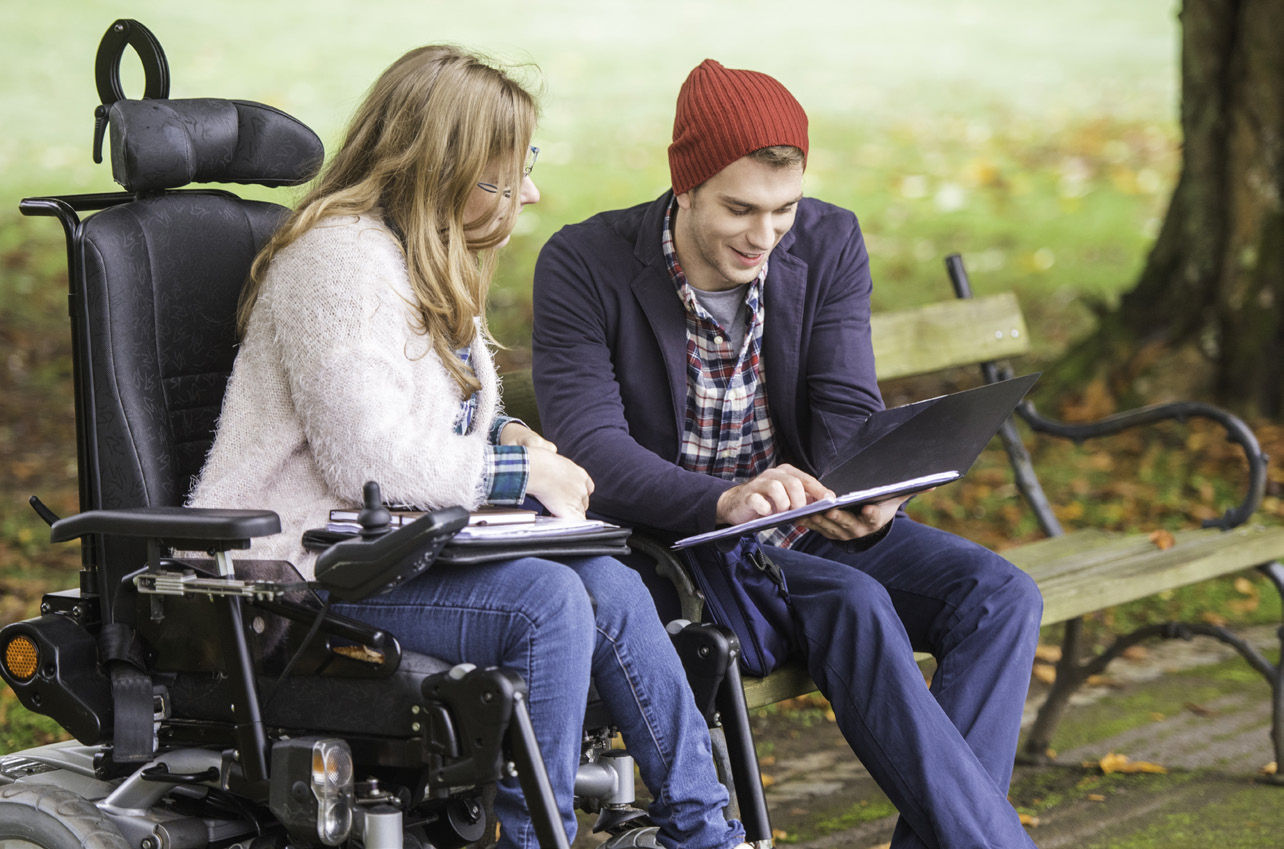 a photograph of a young man showing a young woman who is using a wheelchair something on his tablet