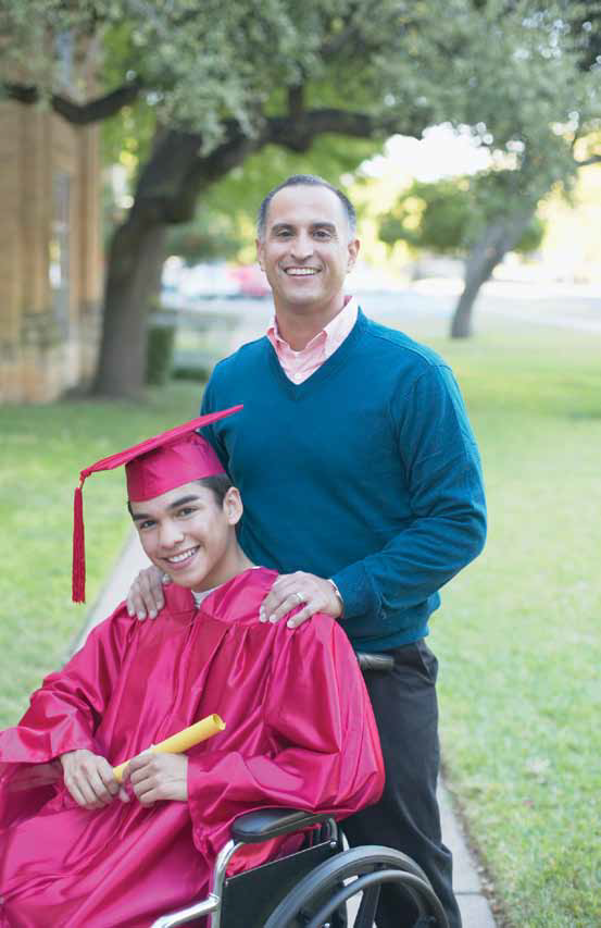 a dad standing proud over his son who is wearing graduation regalia and using a wheelchair