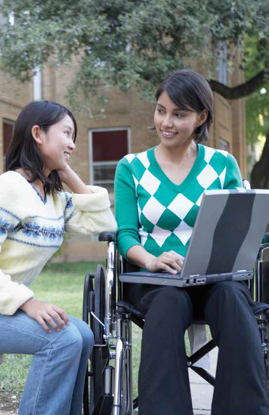 a photo of two female students, one using a wheelchair conversing over something displayed on a laptop