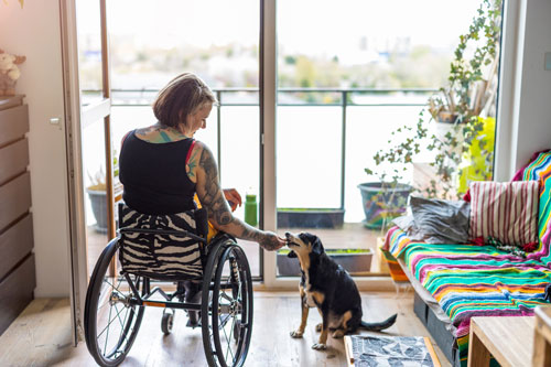 a woman sitting in a wheelchair in her living room, giving her dog a treat. The room has a balcony that overlooks a river