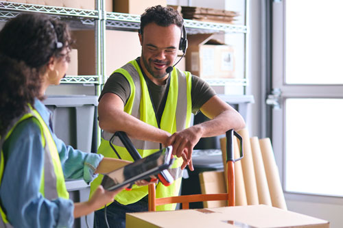 a man with Downs Syndrome wearing a yellow vest and a headset leaning on a dolly on a warehouse floor. He is smiling and talking to a woman who is his coworker holding an iPad