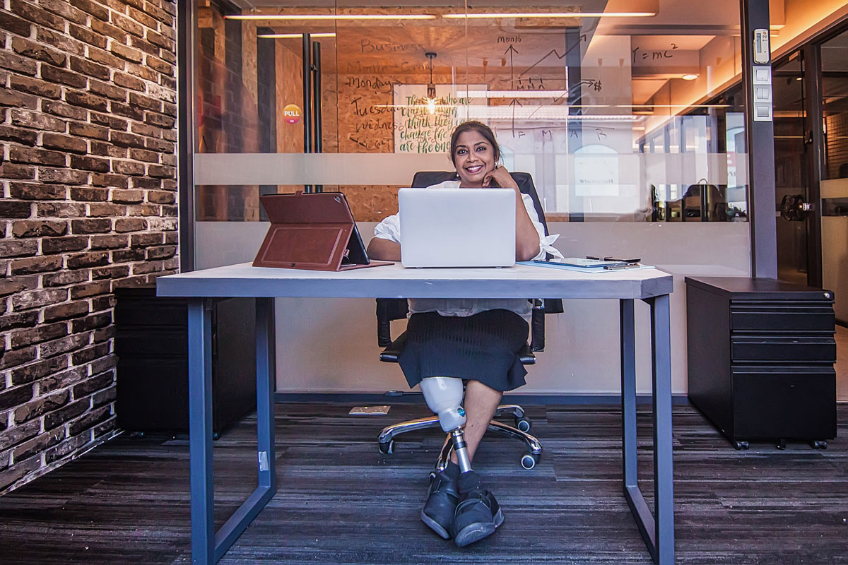 a woman with an artifical leg sitting cross-legged at a desk with a computer and smiling